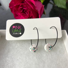 Load image into Gallery viewer, Black hoops with white polymer clay circles on an earring card on a box with a rose behind