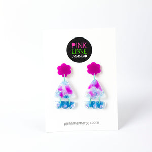Vibrant handcrafted resin earrings with a pink flower top, the middle parts are frosted white triangles with pink cloud patterns and a sprinkling of electric blue glitter. The base element has sparkly lilac flowers perfectly placed above a layer of electric blue glitter. Complimented with mint green jump rings. Earrings on a Pink Lime Mango backing card.
