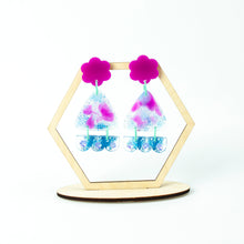 Load image into Gallery viewer, Hexagonal display stand. Vibrant handcrafted resin earrings with a pink flower top, the middle parts are frosted white triangles with pink cloud patterns and a sprinkling of electric blue glitter. The base element has sparkly lilac flowers perfectly placed above a layer of electric blue glitter. Complimented with mint green jump rings.
