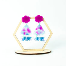 Load image into Gallery viewer, Hexagonal display stand. Vibrant handcrafted resin earrings with a pink flower top, the middle parts are frosted white triangles with pink cloud patterns and a sprinkling of electric blue glitter. The base element has sparkly lilac flowers perfectly placed above a layer of electric blue glitter. Complimented with mint green jump rings.