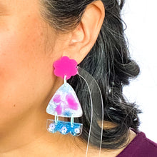 Load image into Gallery viewer, Vibrant handcrafted resin earrings with a pink flower top, the middle parts are frosted white triangles with pink cloud patterns and a sprinkling of electric blue glitter. The base element has sparkly lilac flowers perfectly placed above a layer of electric blue glitter. Complimented with mint green jump rings. Modelled.