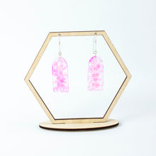 Load image into Gallery viewer, Delicate arches of clear resin with bursts of pale pink and tiny shimmery flower sequins. The unique V shaped earring hooks are made of sterling silver. Displayed on a hexagonal earring display stand.
