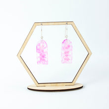 Load image into Gallery viewer, Delicate arches of clear resin with bursts of pale pink and tiny shimmery flower sequins. The unique V shaped earring hooks are made of sterling silver. Displayed on a hexagonal earring display stand.