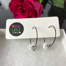 Load image into Gallery viewer, Black hoops with white polymer clay circles on an earring card on a box with a rose behind
