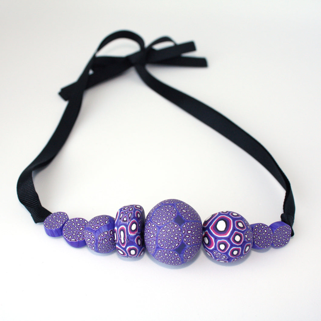 Measurements: each bead is between 1.5cm to 3.5cm Each design is exclusively unique and one of a kind! Finished with a lovely black ribbon tie so you can adjust the necklace length. A beautiful, bold and colourful statement necklace featuring eight different millefiori style handmade beads.  The colours are purple, pink and white.
