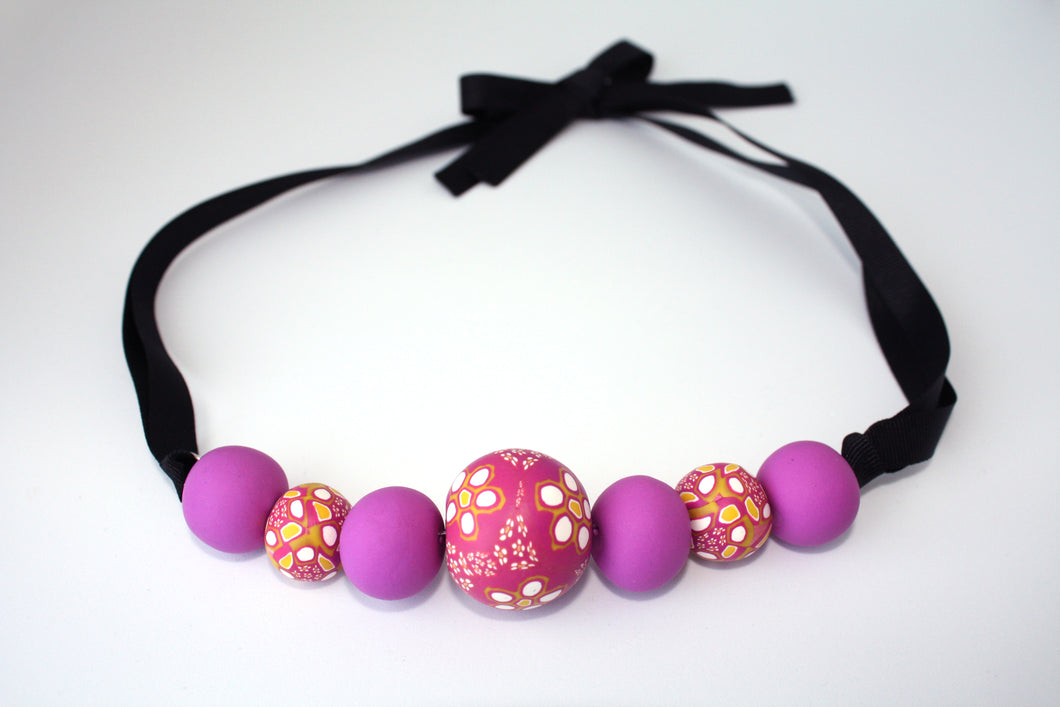 A bold and colourful statement necklace featuring millefiori style handmade beads.  Seven beautiful beads decorated with purples, pinks, white and mustard yellow. With a black ribbon tie closure
