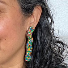 Load image into Gallery viewer, Model wears mosaic rocks squiggle earrings with a beautiful pattern of marbled brown, green, cream, bright red, electric blue, mango orange with a chocolate coloured brown base. 
