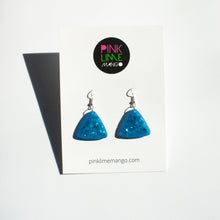 Load image into Gallery viewer, glittery blue resin triangle earrings with silver plated hooks placed on a Pink Lime Mango backing card.
