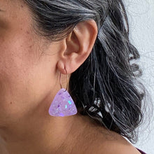 Load image into Gallery viewer, Sparkly lilac triangular earrings from the Under the Sea resin collection. Beautiful layers of fine iridescent glitter, colour shifting aurora flakes and tiny pieces of metallic purple foil. A unique design that has been complimented with multicoloured rainbow hoops! Earrings modelled with a purple top.
