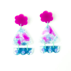 Vibrant handcrafted resin earrings with a pink flower top, the middle parts are frosted white triangles with pink cloud patterns and a sprinkling of electric blue glitter. The base element has sparkly lilac flowers perfectly placed above a layer of electric blue glitter. Complimented with mint green jump rings. Image shows the fronts of the earrings.