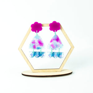 Hexagonal display stand. Vibrant handcrafted resin earrings with a pink flower top, the middle parts are frosted white triangles with pink cloud patterns and a sprinkling of electric blue glitter. The base element has sparkly lilac flowers perfectly placed above a layer of electric blue glitter. Complimented with mint green jump rings.