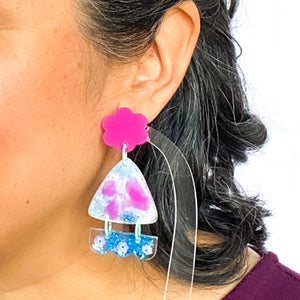 Vibrant handcrafted resin earrings with a pink flower top, the middle parts are frosted white triangles with pink cloud patterns and a sprinkling of electric blue glitter. The base element has sparkly lilac flowers perfectly placed above a layer of electric blue glitter. Complimented with mint green jump rings. Modelled.