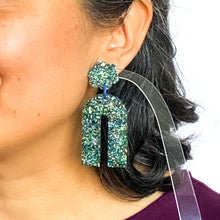 Load image into Gallery viewer, Model shot. Handcrafted resin arch earrings that are jam packed with sparkly teal blue, green and gold glitter! These beauties have flower stud tops and look fabulous on!
