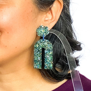 Model shot. Handcrafted resin arch earrings that are jam packed with sparkly teal blue, green and gold glitter! These beauties have flower stud tops and look fabulous on!